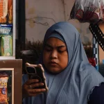 Perempuan berkerudung memegang ponsel di samping rak jajanan warung kecil