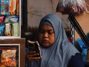 Perempuan berkerudung memegang ponsel di samping rak jajanan warung kecil