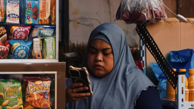 Perempuan berkerudung memegang ponsel di samping rak jajanan warung kecil