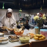Perempuan berkerudung menata makanan di meja makan penuh hidangan di dalam rumah