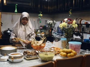 Perempuan berkerudung menata makanan di meja makan penuh hidangan di dalam rumah