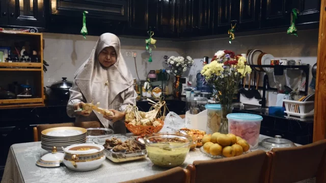 Perempuan berkerudung menata makanan di meja makan penuh hidangan di dalam rumah