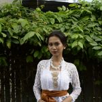 Ilustrasi seorang perempuan berkebaya putih berdiri di antara dedaunan hijau.