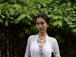 Ilustrasi seorang perempuan berkebaya putih berdiri di antara dedaunan hijau.