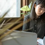 Perempuan muda membaca lembar berkas di depan komputer jinjing di meja kedai kopi