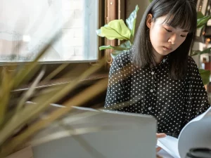 Perempuan muda membaca lembar berkas di depan komputer jinjing di meja kedai kopi