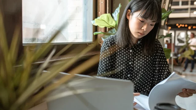 Perempuan muda membaca lembar berkas di depan komputer jinjing di meja kedai kopi