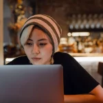 Perempuan berkupluk garis menatap layar laptop di meja sebuah kafe