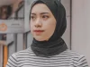 Perempuan muda berhijab hitam dan sweter bergaris berdiri di tepi jalan perkotaan