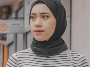 Perempuan muda berhijab hitam dan sweter bergaris berdiri di tepi jalan perkotaan