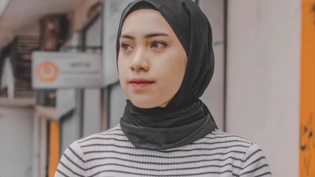 Perempuan muda berhijab hitam dan sweter bergaris berdiri di tepi jalan perkotaan