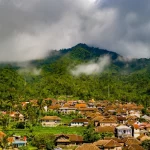 Permukiman desa beratap genting di kaki perbukitan berhutan yang berkabut