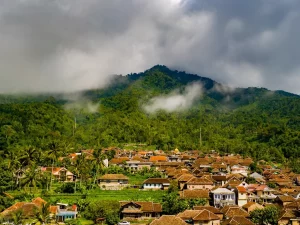 Permukiman desa beratap genting di kaki perbukitan berhutan yang berkabut