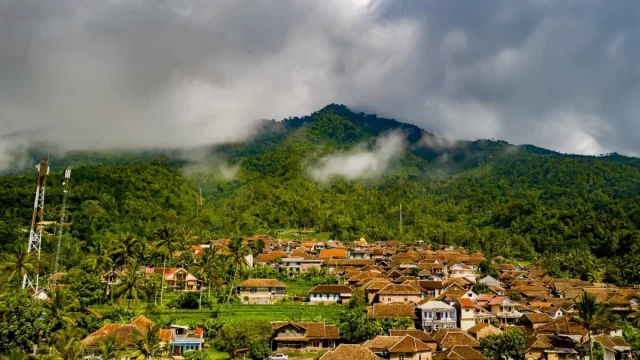 Permukiman desa beratap genting di kaki perbukitan berhutan yang berkabut