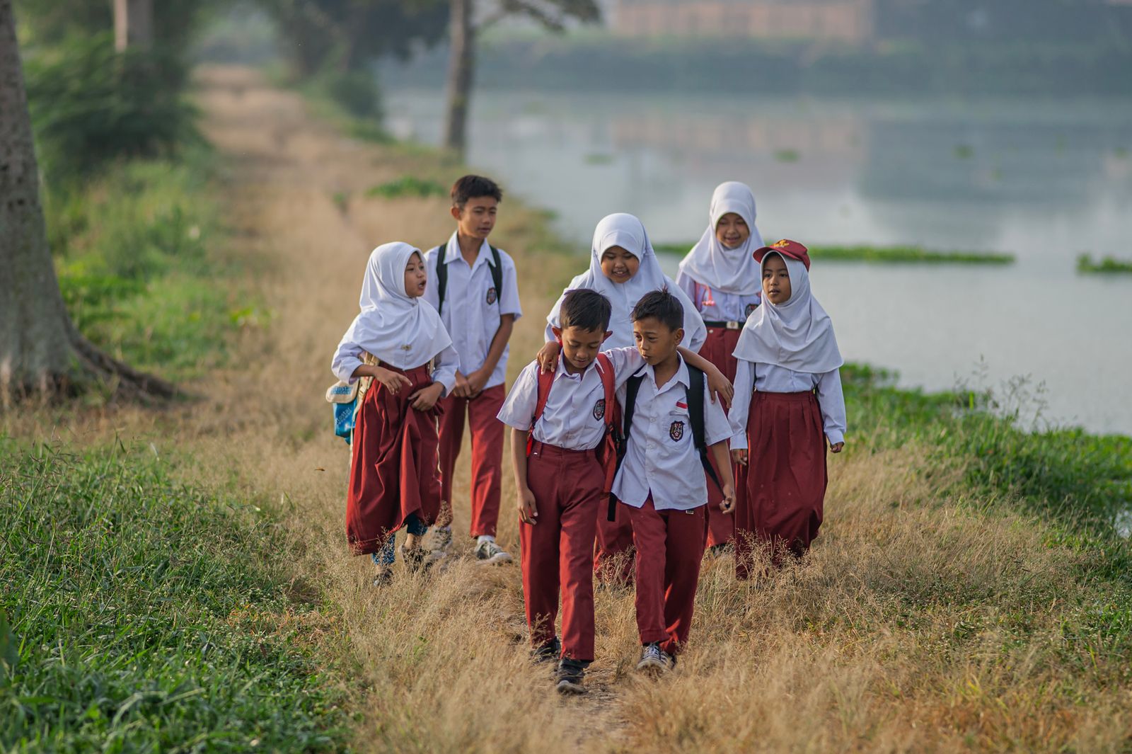 Ilustrasi sekelompok murid sekolah dasar berseragam merah putih berjalan beriringan menyusuri jalan setapak berumput di tepi air.