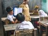 Ilustrasi seorang guru berdiri di dekat papan tulis hitam sementara murid-murid berseragam duduk di bangku kayu ruang kelas sederhana.