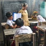 Ilustrasi seorang guru berdiri di dekat papan tulis hitam sementara murid-murid berseragam duduk di bangku kayu ruang kelas sederhana.
