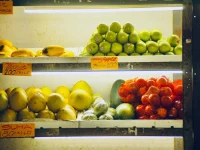 Rak toko berisi pisang, jambu, jeruk bali, sayur, dan tomat berlabel harga per kilo
