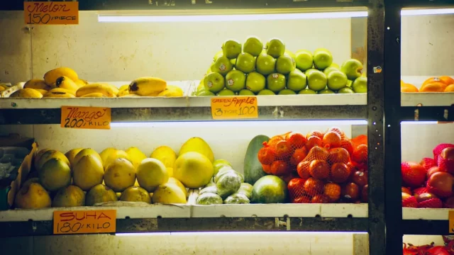 Rak toko berisi pisang, jambu, jeruk bali, sayur, dan tomat berlabel harga per kilo