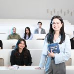 Ilustrasi seorang mahasiswa berdiri memegang buku di ruang kuliah bersama teman-teman sekelasnya.
