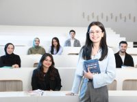 Ilustrasi seorang mahasiswa berdiri memegang buku di ruang kuliah bersama teman-teman sekelasnya.