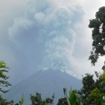 Ilustrasi gunung api menyemburkan kolom abu tebal dilihat dari balik pepohonan.