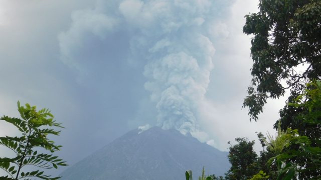 Ilustrasi gunung api menyemburkan kolom abu tebal dilihat dari balik pepohonan.