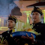 Ilustrasi dua lelaki berbusana Jawa membawa sesaji di tengah kepulan asap dupa.