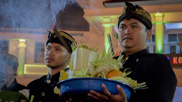 Ilustrasi dua lelaki berbusana Jawa membawa sesaji di tengah kepulan asap dupa.