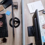 Dua pekerja mengetik di papan ketik komputer pada meja kantor bersekat