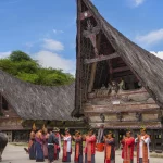 Rumah Bolon beratap menjulang dengan rombongan berbusana adat di halamannya