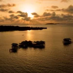 Rumah panggung di atas laut dengan latar pulau saat matahari terbenam