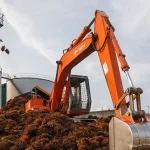 Ekskavator oranye mengeruk tumpukan tandan kelapa sawit di halaman pabrik sawit