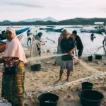 Ilustrasi warga membersihkan jaring dan memilah hasil tangkapan di tepi pantai tempat perahu nelayan bersandar