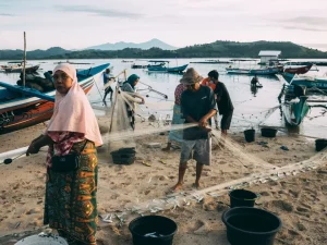 Ilustrasi warga membersihkan jaring dan memilah hasil tangkapan di tepi pantai tempat perahu nelayan bersandar