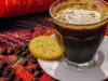 Segelas kopi hitam berbusa di samping biji kopi sangrai di atas kain bermotif merah