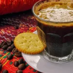Segelas kopi hitam berbusa di samping biji kopi sangrai di atas kain bermotif merah