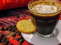Segelas kopi hitam berbusa di samping biji kopi sangrai di atas kain bermotif merah