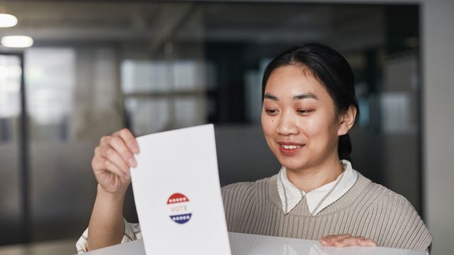 Ilustrasi seorang perempuan memegang surat suara di ruang pemungutan suara.