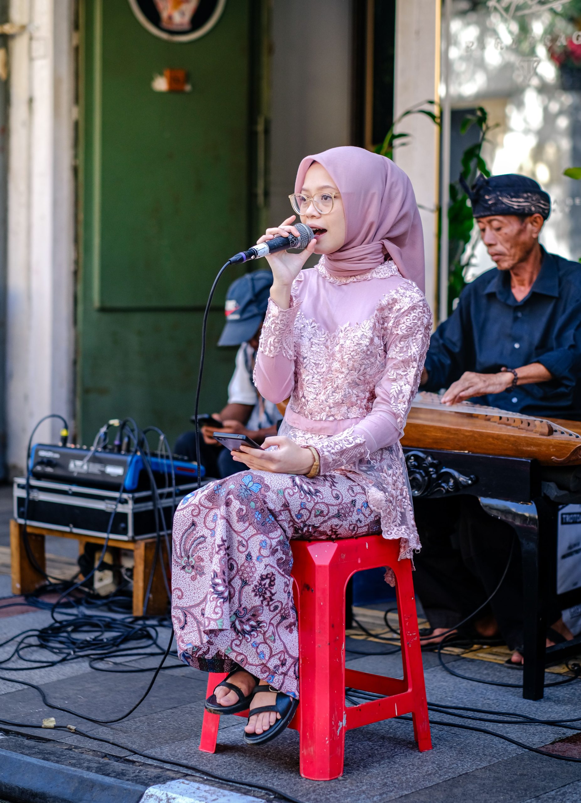 Ilustrasi perempuan berbusana merah muda duduk di kursi kecil membawakan lagu diiringi pemusik di belakangnya.