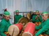 Ilustrasi kelompok pemusik tradisi berbusana hijau memainkan gendang dalam sebuah pertunjukan.