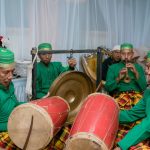 Ilustrasi kelompok pemusik tradisi berbusana hijau memainkan gendang dalam sebuah pertunjukan.