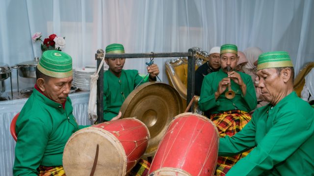 Ilustrasi kelompok pemusik tradisi berbusana hijau memainkan gendang dalam sebuah pertunjukan.