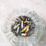 Ilustrasi asbak kaca penuh puntung rokok dan abu.