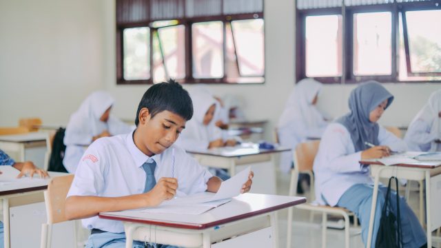 Ilustrasi murid-murid duduk di bangku kelas mengerjakan tugas tulis.