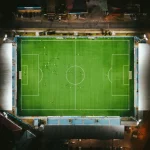 Stadion sepak bola benderang dengan pemain di lapangan dilihat dari udara pada malam hari