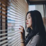 Ilustrasi seorang perempuan menatap ke luar melalui bilah tirai jendela.