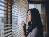 Ilustrasi seorang perempuan menatap ke luar melalui bilah tirai jendela.