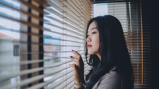Ilustrasi seorang perempuan menatap ke luar melalui bilah tirai jendela.