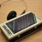 Ilustrasi pemutar kaset jinjing lawas beserta headphone di atas permukaan kayu.